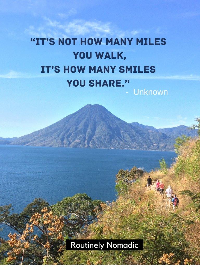 Line of 5 people hiking on trail on hill with lake and volcano in background and a hiking with friends quotes on top